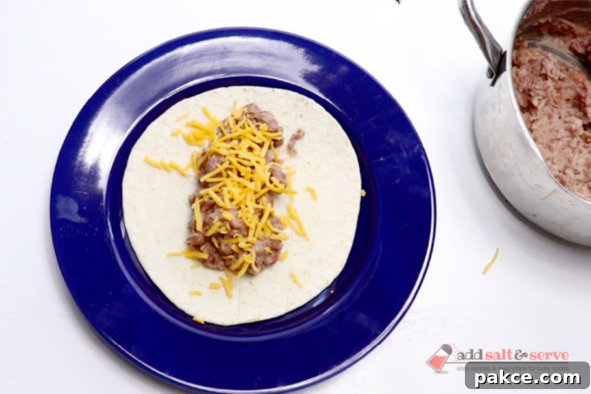 Classic Smothered Burritos 8 A warm flour tortilla with a line of bean burrito filling, topped with a generous sprinkle of shredded cheddar cheese, ready for folding.