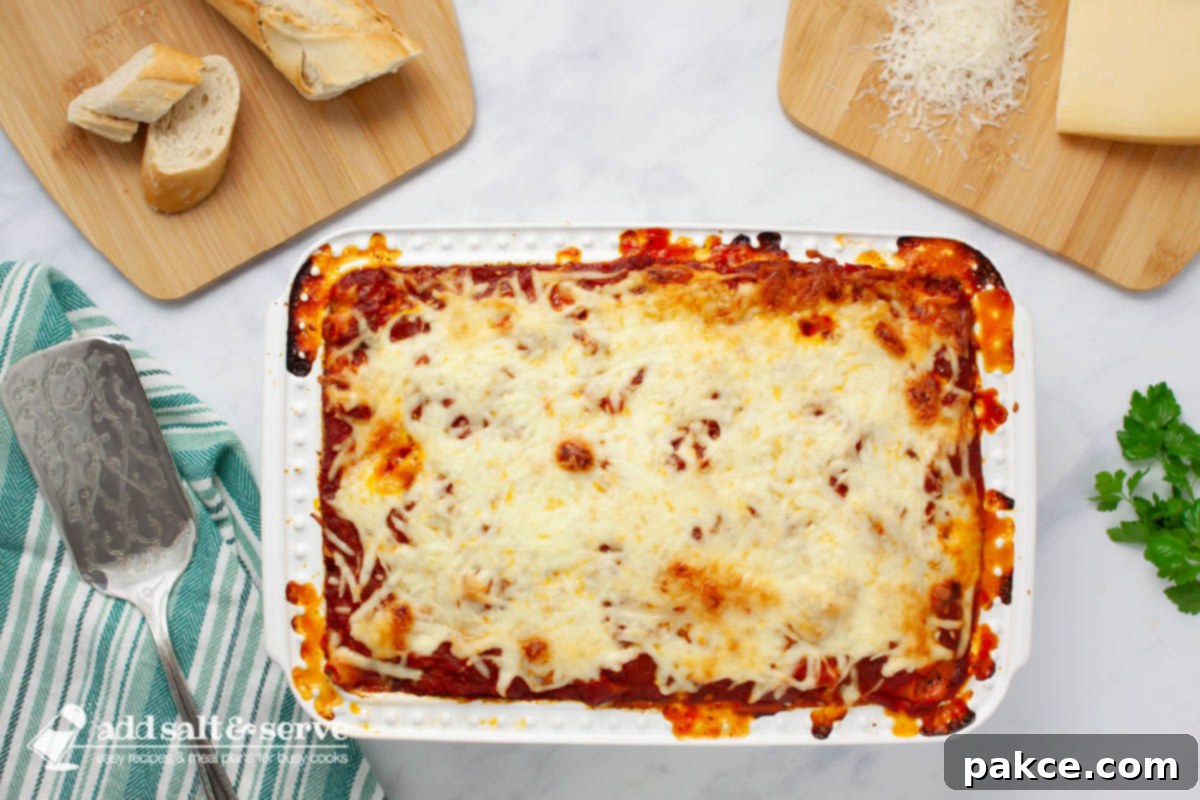 Classic Cheesy Meatless Baked Ziti 8 Pan of cheesy baked ziti on a counter surrounded by sprigs of fresh parsley, sliced baguette, and Parmesan cheese.