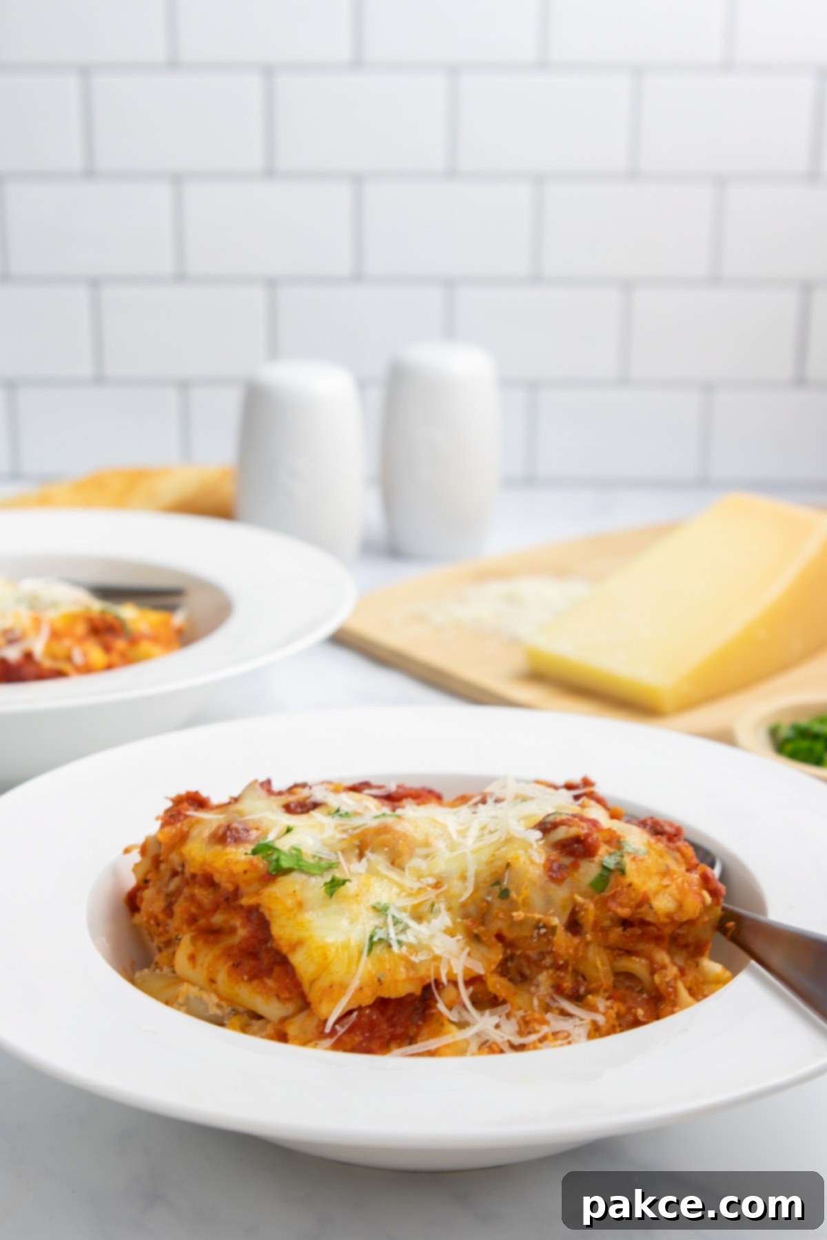 Classic Cheesy Meatless Baked Ziti 9 A serving of cheesy meatless baked ziti in a bowl with a fork with a cutting board with parmesan cheese in the background.