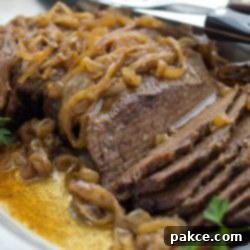 Tender Crockpot London Broil Au Jus 9 Slices of tender beef on a white platter, covered with cooked onions and resting in a rich au jus. The dish is garnished with fresh parsley and a steak knife rests on the platter's edge.