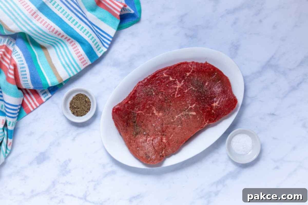 Tender Crockpot London Broil Au Jus 4 Uncooked London broil roast seasoned generously with salt and pepper on a cutting board.