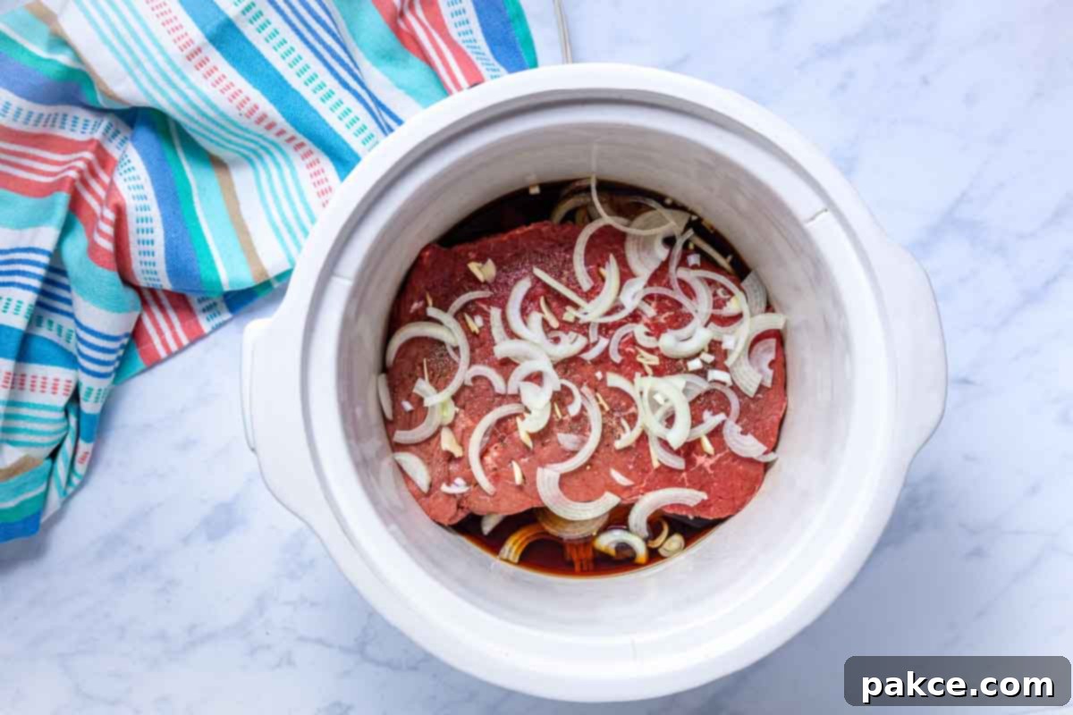 Tender Crockpot London Broil Au Jus 7 A crockpot showing layers of sliced onions, seasoned uncooked London broil roast, and liquid ingredients (soy sauce and water) with remaining onions on top.
