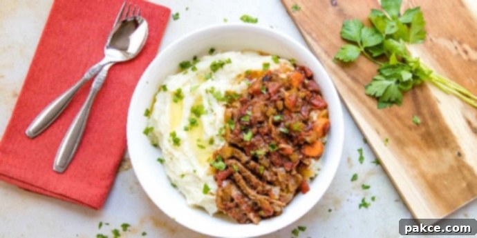White bowl with mashed potatoes in one half and strips of beef and chopped vegetables in the other half, garnished with parsley. To the left of the bowl is a spoon and fork on a red linen napkin. To the right is a wooden cutting board with parsley on it.