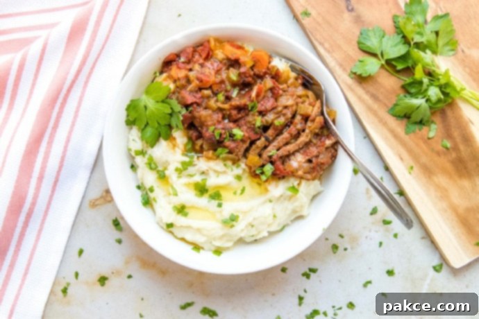 White bowl with mashed potatoes in one half and strips of beef and chopped vegetables in the other half, garnished with parsley. To the left of the bowl is a spoon and fork on a red linen napkin. To the right is a wooden cutting board with parsley on it.