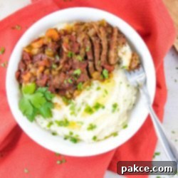 Perfect Swiss Steak: Slow Cooker or Instant Pot 2 White bowl with mashed potatoes in one half and strips of beef and chopped vegetables in the other half, garnished with parsley. The bowl is on a red linen napkin and there is a fork in the bowl.