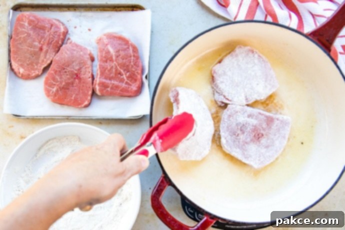 Three floured beef steaks in a red and white skillet. To the left of the skillet are three plain beef steaks on a metal pan and a white plate with the flour mixture.
