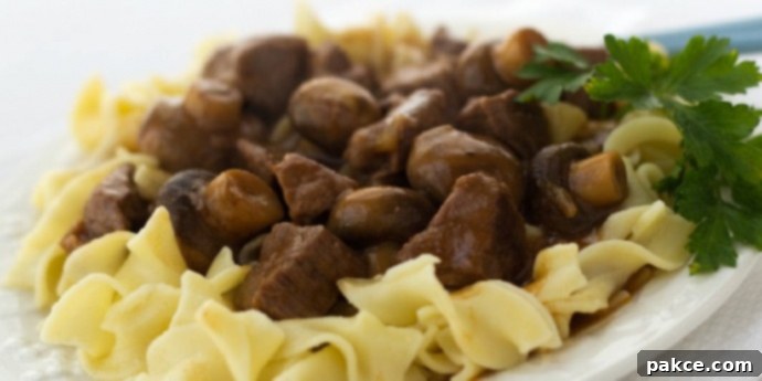 A white plate showcasing a hearty portion of Slow Cooker Beef and Mushrooms served over fluffy egg noodles, garnished with fresh parsley. This comforting meal is perfect for busy evenings and family dinners.