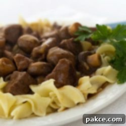 Hearty Slow Cooker Beef and Mushrooms 2 White plate with egg noodles topped with beef and mushrooms, garnished with a sprig of parsley.