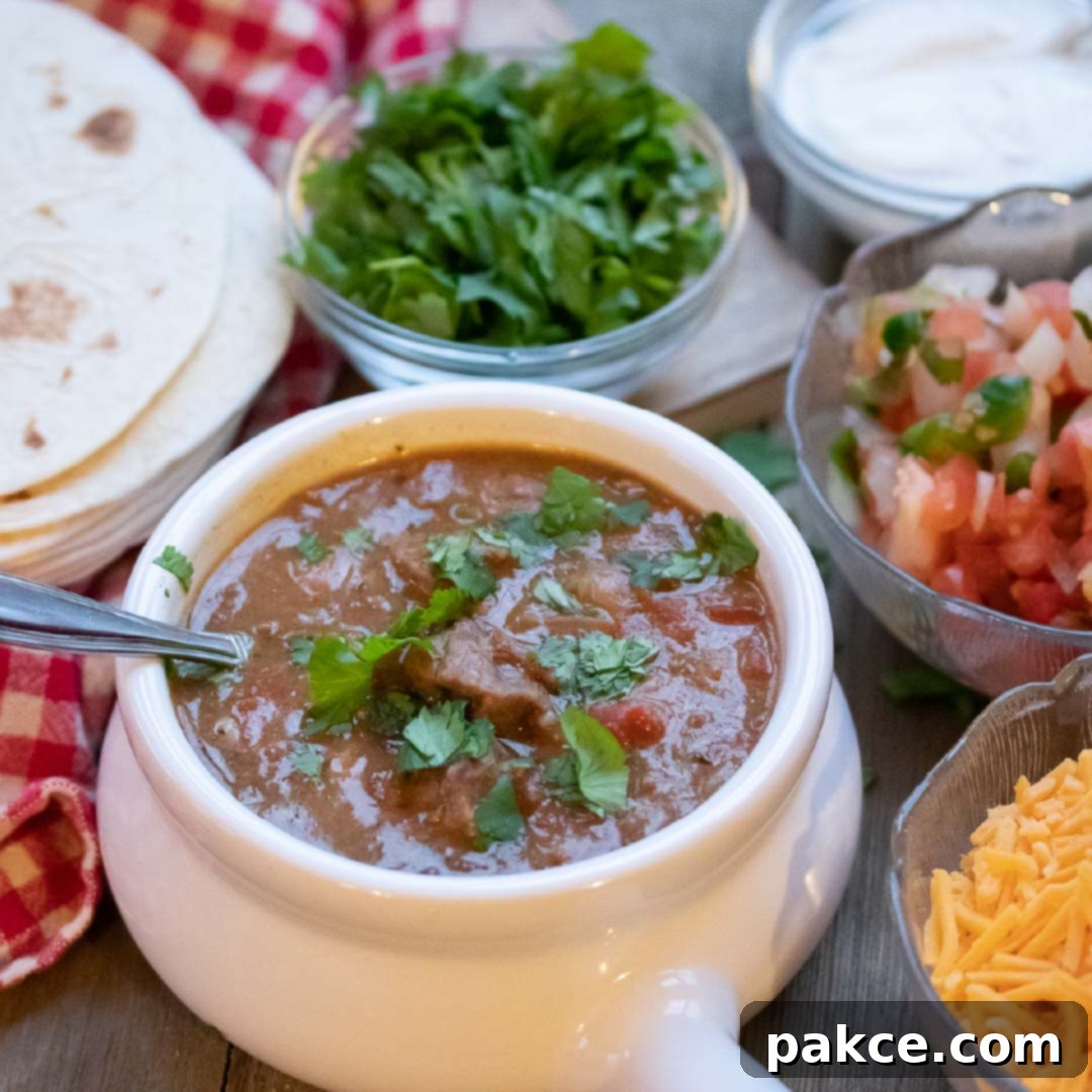 Carne Guisada (Mexican Beef Stew) in a white crock with cheese, tortillas, fresh cilantro, sour cream, and pico de gallo