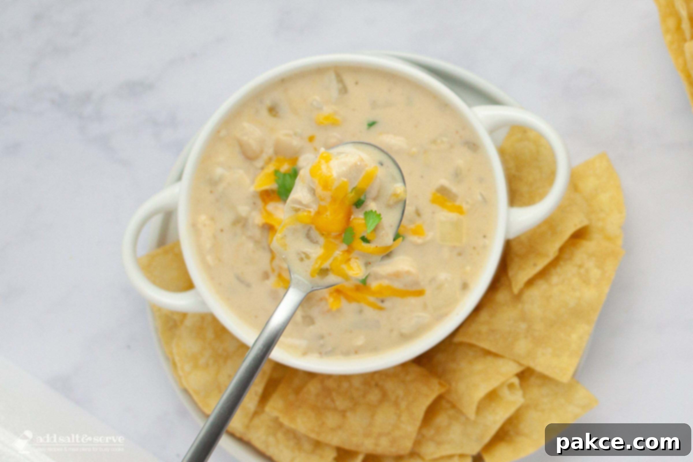 30-Minute Stovetop Creamy White Chicken Chili 7 A bowl of creamy white chicken chili garnished with cheddar cheese and cilantro and served with chips with a spoonful held above the bowl toward the camera.