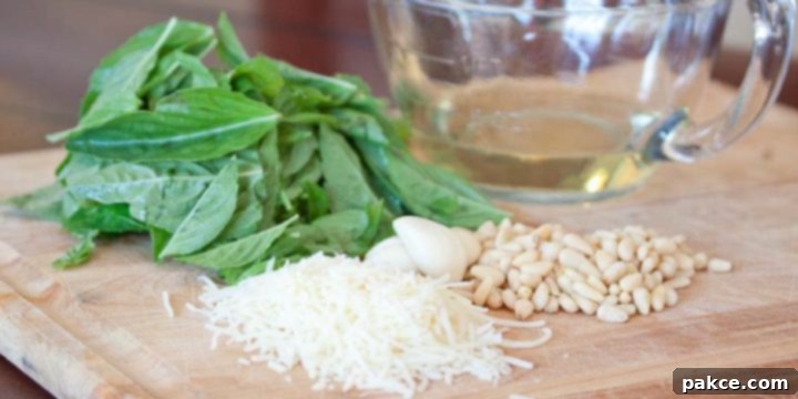Ingredients for making homemade pesto: a pile of fresh basil leaves, a glass measuring cup with olive oil, a bowl of shredded Parmesan cheese, several garlic cloves, and a pile of pine nuts, all arranged on a wooden cutting board.