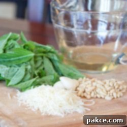 DIY Pesto Perfection 2 Close-up photo of all the raw ingredients for homemade pesto beautifully arranged on a wooden board: fresh basil leaves, olive oil in a glass measuring cup, shredded Parmesan cheese, garlic cloves, and pine nuts.