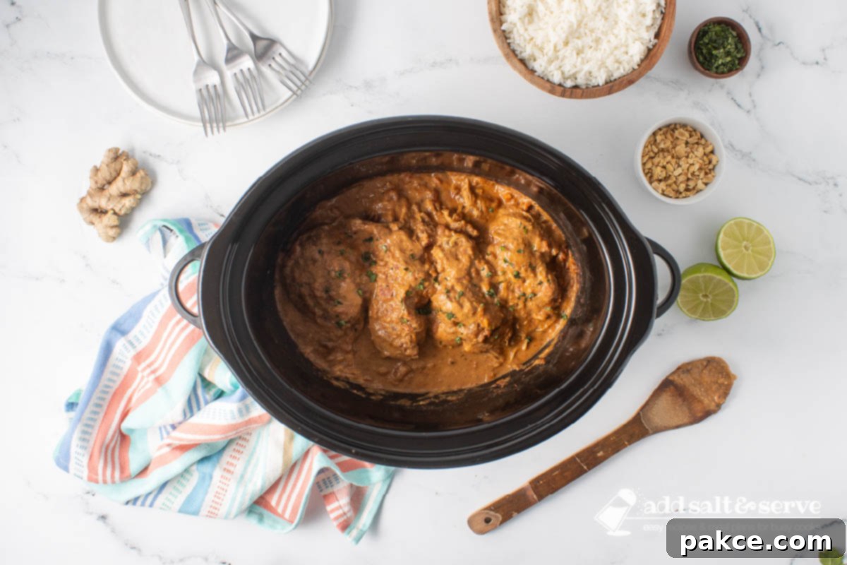 Creamy Slow Cooker Thai Peanut Chicken 7 Cooked Thai Peanut Chicken in a slow cooker surrounded by rice, chopped peanuts, cilantro, ginger, a halved lime, and a stack of empty plates with forks.