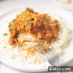 Creamy Slow Cooker Thai Peanut Chicken 9 Plated slow cooker Thai Peanut Chicken over rice garnished with chopped peanuts and cilantro with a fork cutting into the chicken showing white breast meat.