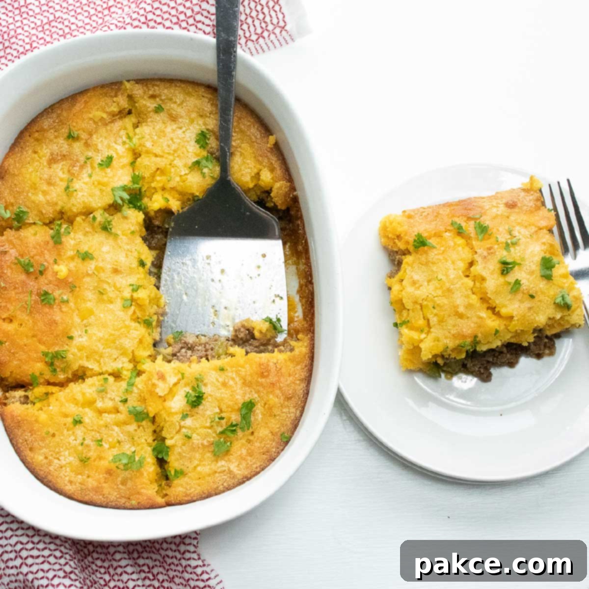Casserole Burger Bake in a white casserole dish. One piece is cut out and sitting on a white plate next to the casserole. There is a spatula in the casserole dish and a fork on the plate.