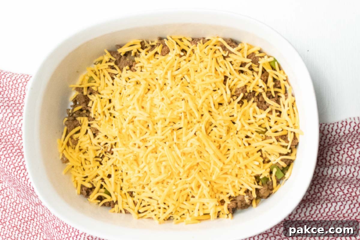 Hearty Cornbread Burger Bake 4 Cooked ground beef, diced onions and bell peppers in a white casserole dish, topped with shredded cheddar cheese.