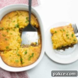 Hearty Cornbread Burger Bake 6 Casserole Burger Bake in a white casserole dish. One piece is cut out and sitting on a white plate next to the casserole. There is a spatula in the casserole dish and a fork on the plate.
