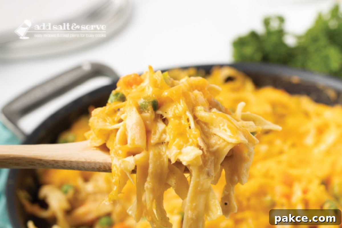 Golden Cheesy Tuna Delight 7 A generous spoonful of creamy cheesy tuna noodle casserole being lifted from a skillet, showcasing the tender noodles, peas, carrots, and rich sauce.