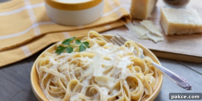 Yellow plate with fettuccine and Alfredo sauce with pot of Alfredo sauce, hunk of Parmesan cheese, and nutmeg grinder in background
