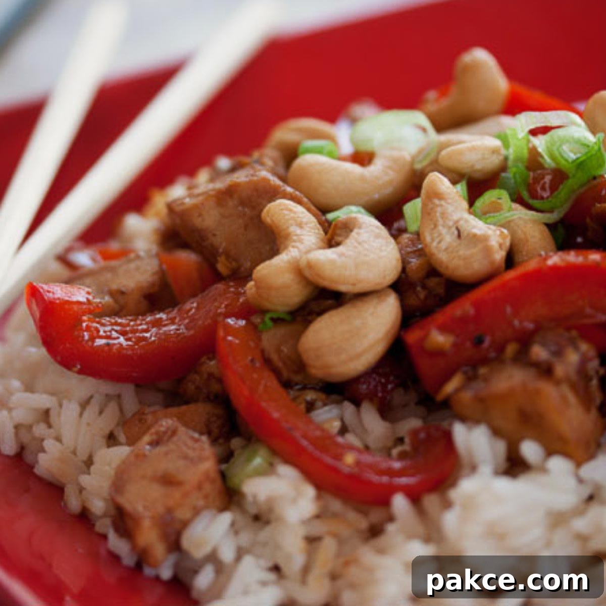 Red plate with chopsticks and rice, sliced red bell peppers, cashews, chicken, and green onions – a vibrant and delicious Chinese Cashew Chicken dish.