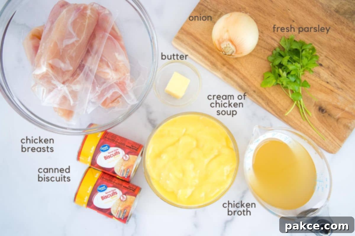 Easy Crockpot Chicken & Dumplings Individually wrapped chicken breasts in a clear glass bowl, two tablespoon butter in a small clear glass bowl, two cans of refrigerated biscuits, cream of chicken soup in a clear glass bowl, chicken broth in a clear measuring cup, and one onion and some sprigs of parsley on a cutting board.