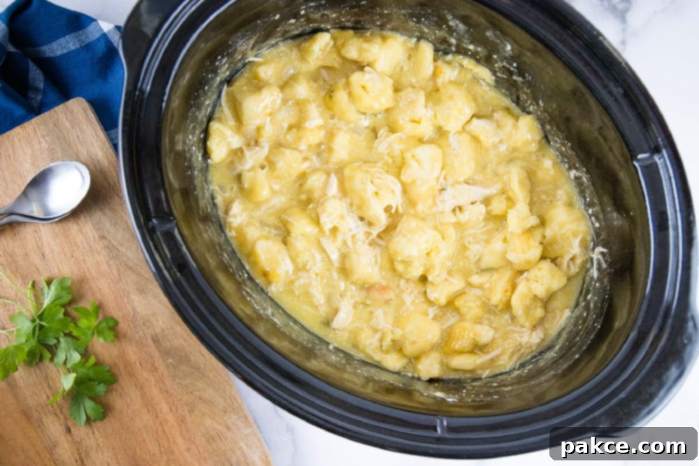 Easy Crockpot Chicken & Dumplings Cooked chicken and dumplings in a slow cooker. Next to the slow cooker is a wooden cutting board with a spoon and sprigs of parsley.