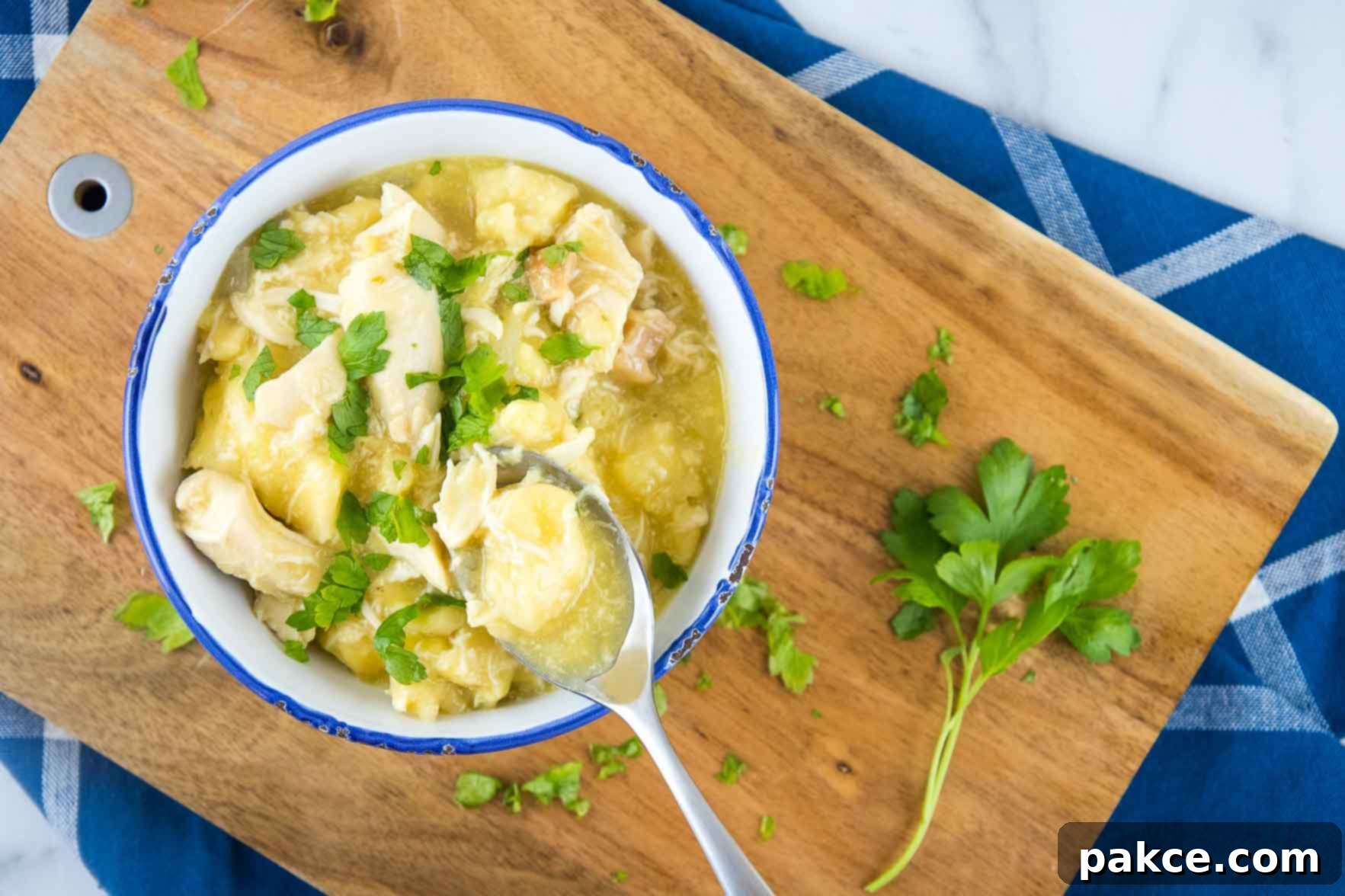 Easy Slow Cooker Chicken and Dumplings with Biscuits 3 Chicken and dumplings garnished with parsley in a white bowl with a blue rim. There is a spoon with a dumpling and some broth. The bowl is on a wooden cutting board on top of a blue napkin with white stripes, all on a white marble counter.