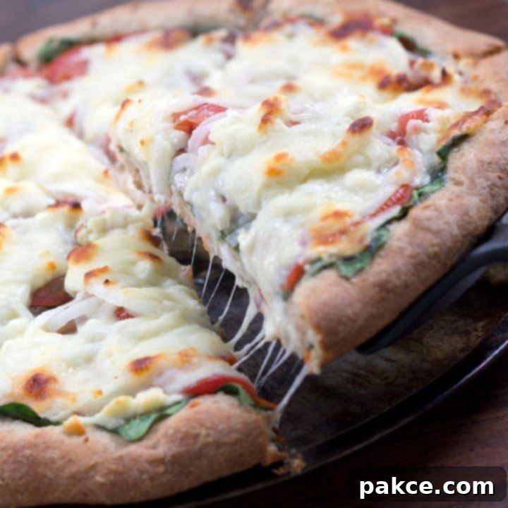 Serving spatula lifting a slice of pizza off of whole pizza on a pizza pan, pizza has thick crust, baby spinach onions, tomatoes, and mozzarella cheese.