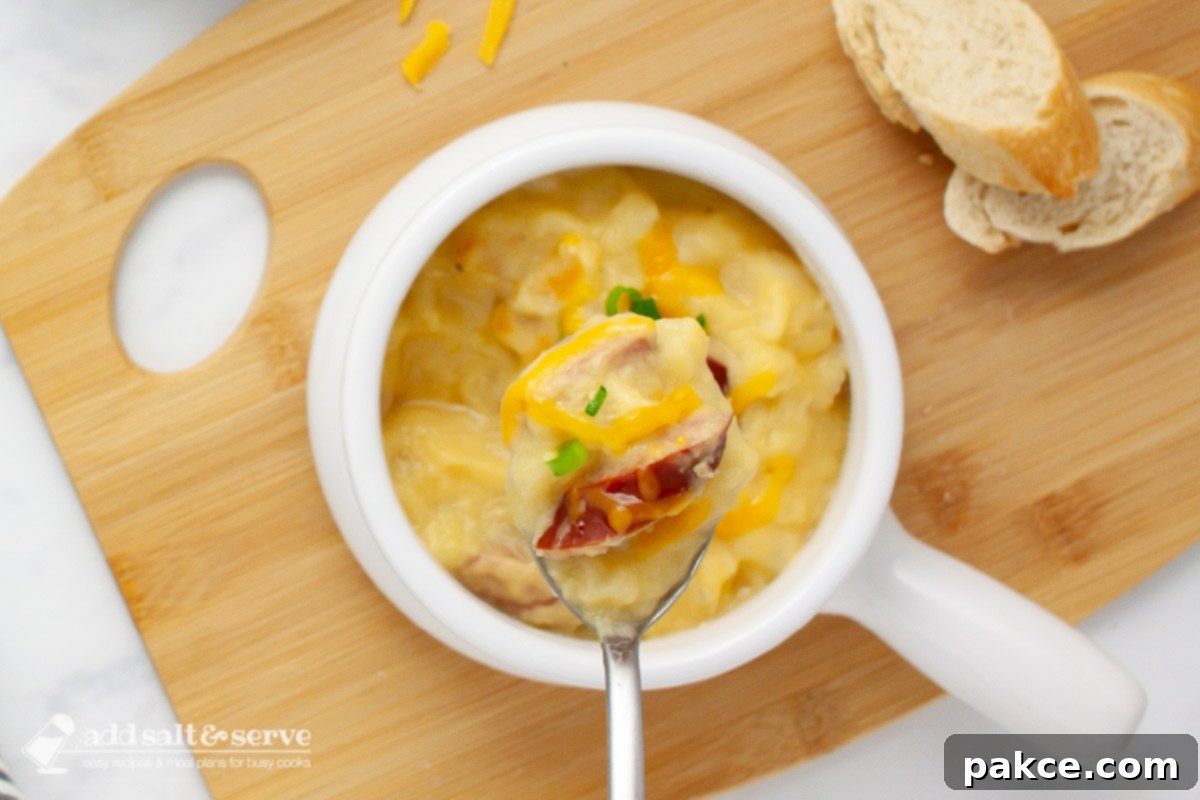 Close-up of a spoonful of cheesy potato soup with kielbasa pieces garnished with shredded cheddar and sliced green onion on a spoon held over a crock of the soup.