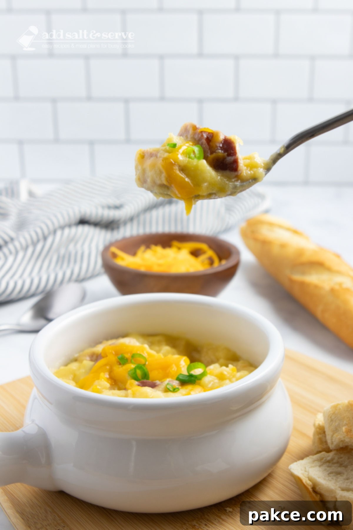 Slow Cooker Cheesy Kielbasa and Potato Soup 6 Crock of cheesy potato soup garnished with shredded cheddar cheese and sliced green onion with a spoonful held above the crock with a baguette in the background.