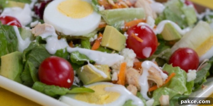 Cobb salad on a white square plate on a yellow tablecloth. Cobb salad ingredients are lettuce, grape tomatoes, sliced boiled eggs, diced avocado, diced chicken, shredded carrots, and blue cheese dressing. A fork is next to the plate, inviting a taste.