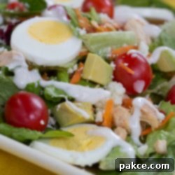 Hearty Chicken Avocado Cobb Salad 2 Cobb salad on a white square plate on a yellow tablecloth. Cobb salad ingredients are lettuce, grape tomatoes, sliced boiled eggs, diced avocado, diced chicken, shredded carrots, and blue cheese dressing.