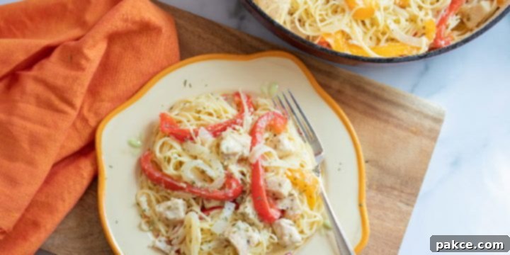 A close-up of creamy chicken pepper pasta on a white plate with a yellow rim. It features angel hair pasta tossed with diced chicken, strips of red and yellow bell peppers, and a rich white sauce. In the background, part of a skillet with more pasta and ingredients is visible on a white marble counter with an orange napkin.