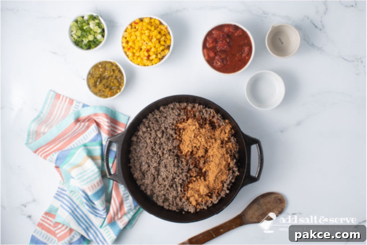 Zesty Mexican Pasta 5 A cast iron skillet with cooked ground beef and taco seasoning blended with water, surrounded by bowls of Mexi-corn, green chiles, fire-roasted diced tomatoes, and chopped green onions, with a wooden spoon.