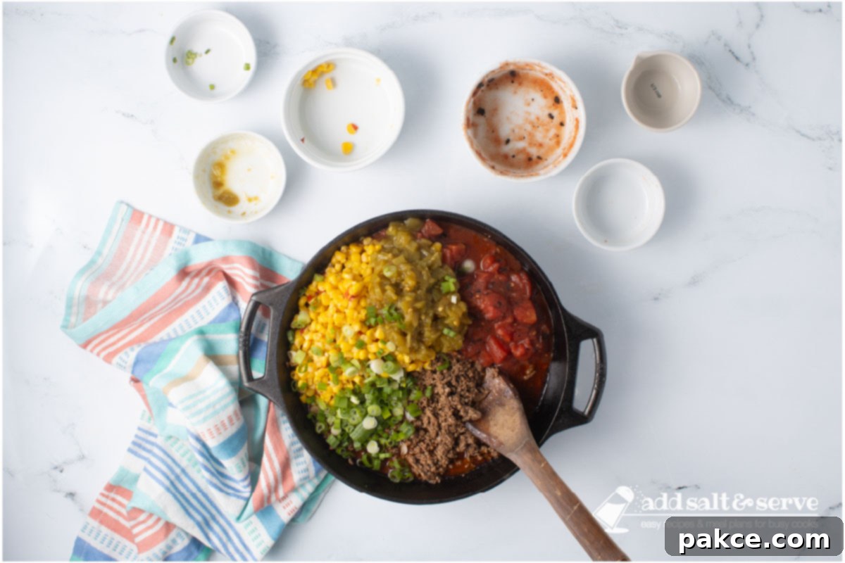 Zesty Mexican Pasta 6 A cast iron skillet containing the fully assembled Mexican Spaghetti sauce: seasoned ground beef, Mexi-corn, green chiles, fire-roasted diced tomatoes, and chopped green onions, all mixed with a wooden spoon.