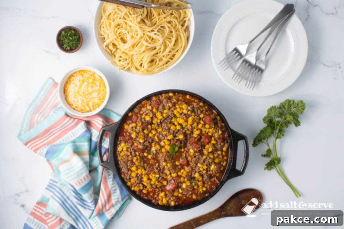 Zesty Mexican Pasta 7 A cast iron skillet filled with simmering Mexican Spaghetti sauce, a bowl of shredded cheddar cheese, and a bowl of cooked spaghetti, all arranged on a kitchen counter with fresh parsley accents.