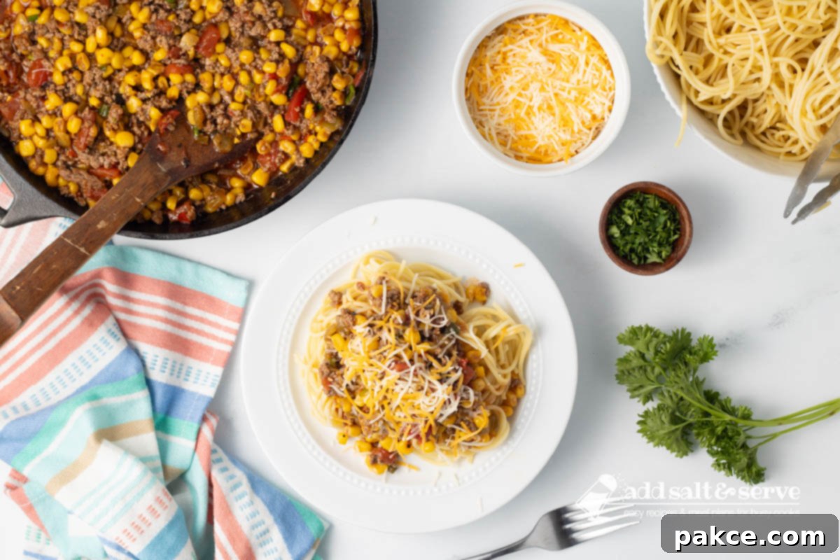 Zesty Mexican Pasta 8 A beautifully plated dish of Mexican Spaghetti topped generously with shredded cheese, next to a serving bowl of cooked spaghetti, a pot of the rich Mexican Spaghetti sauce, and a dish of extra shredded cheddar cheese. Fresh tomatoes and parsley add a decorative touch to the kitchen counter.