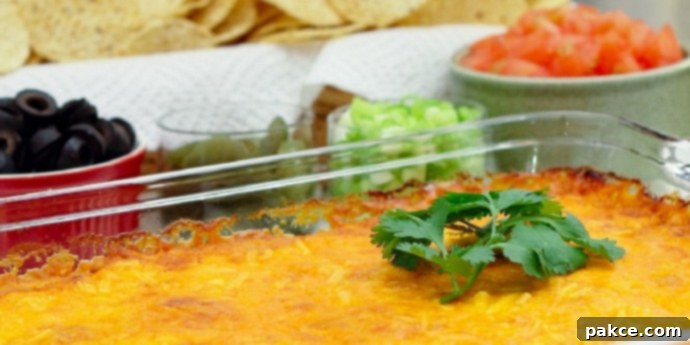 Clear glass casserole dish with beans topped with melted cheese and garnished with a sprig of cilantro. In the background is a red bowl of sliced black olives, a clear bowl of jalepeno slices, a clear bowl of diced green onions, a white bowl of diced tomatoes, and a basket of tortilla chips.