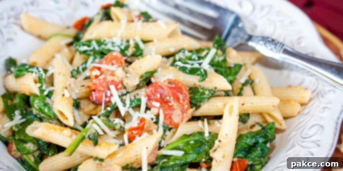 White plate with penne pasta, cooked baby spinach, cherry tomatoes, and shredded mozzarella cheese. There is a fork on the plate.