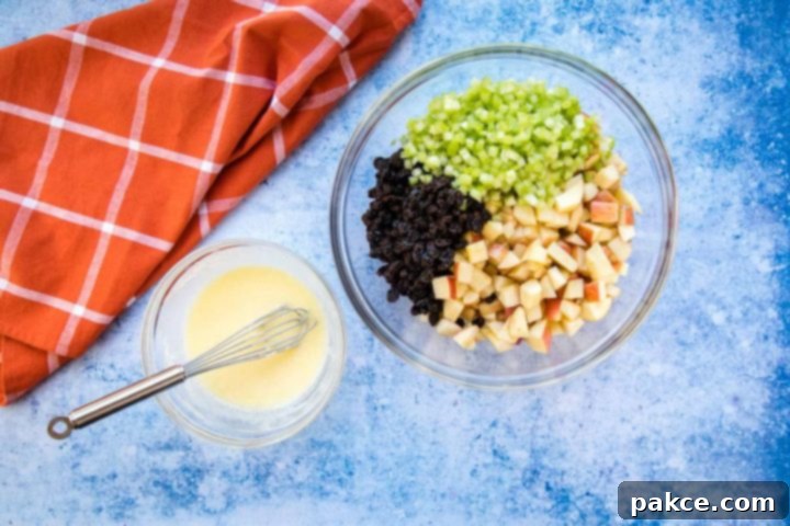 apple-raisin-salad-ip-2-min Overhead view of a large bowl with chopped apples, raisins, and celery, a smaller bowl with a creamy salad dressing and a whisk, all on a blue surface with a coral plaid towel to the side