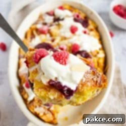 French toast bake with berries and almonds garnished with powdered sugar, whipped cream, and jam in a casserole dish with a serving held toward the camera.